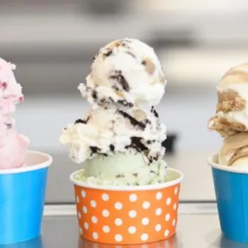 Three cups of ice cream with two scoops each, featuring different flavors, are lined up on a counter against an orange background.