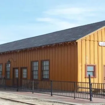 A yellow wooden train station building labeled "CAMPO" with a signal post nearby, surrounded by a black metal fence on a clear day.