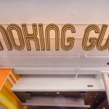 Large gold letters spelling "SMOKING GUN" are mounted on the ceiling inside a modern, brightly lit building.