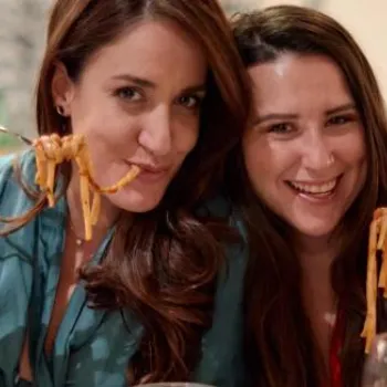 Two women sit at a table in a restaurant, smiling at the camera while holding forks with pasta.
