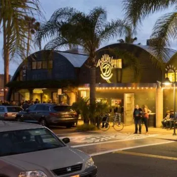 People stand outside a lit-up building with arched roofs and palm trees at dusk, while cars are parked along the street in front.