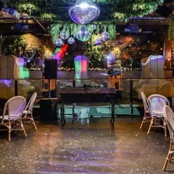 A nightclub interior with scattered chairs and tables, hanging disco balls, plants, and colorful lighting, creating a lively and modern atmosphere.