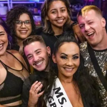 A group of eight people pose closely together in a lively indoor setting; one person in the center wears a sash labeled "MS." and everyone is smiling.
