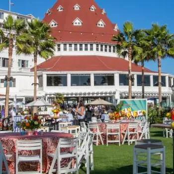 Outdoor event setup with round tables and chairs on a grassy area in front of a large historic hotel with a red-roofed turret, palm trees, and clear blue sky.