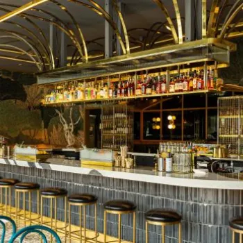 Modern bar with a marble countertop, gold accents, high stools, and shelves of liquor bottles, set against a leafy-patterned wallpaper and decorative lighting.
