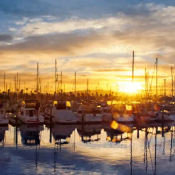 Sunset over a marina with numerous docked boats and sailboats; the sky is partly cloudy, and the water reflects the colors of the setting sun.