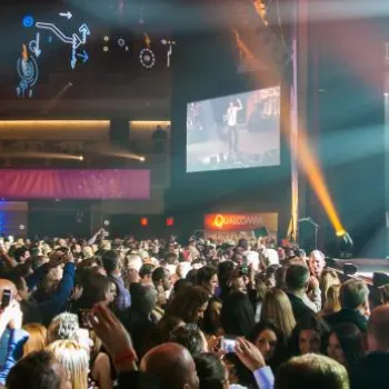 A large crowd watches a live band perform on stage at an indoor venue with bright stage lights and a screen displaying the performance.