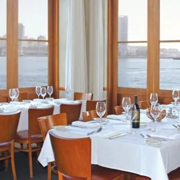 A dining area with tables set for a meal, wooden chairs, white tablecloths, large windows overlooking a waterfront cityscape, and a wall mirror reflecting the room.