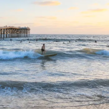 Several people surf near a wooden pier at sunset, with waves breaking onto the sandy shore and scattered clouds in the sky.