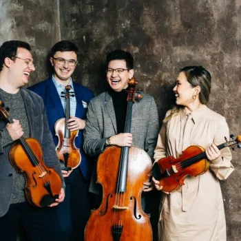 Four musicians stand together holding string instruments, smiling and talking; two have violins, one has a viola, and one holds a cello, against a textured background.