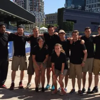 A group of eleven people in matching uniforms pose together on a baseball field near the stands, with buildings visible in the background.