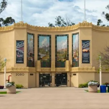 The exterior of the San Diego Automotive Museum with art deco architecture, banners, outdoor seating, and colorful umbrellas in the foreground.