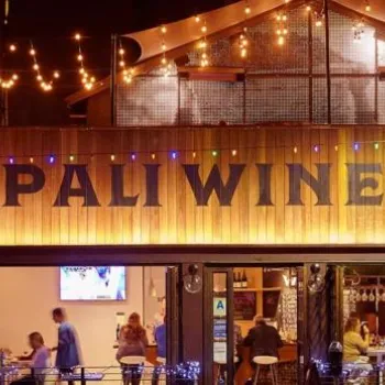 Outdoor view of Pali Wine Co. at night, with string lights above, people sitting at tables inside and outside, and a wooden facade featuring the company’s name.