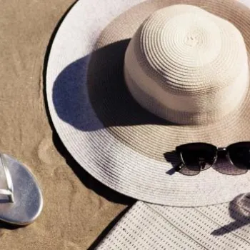 A sun hat, sunglasses, sandals, and a drink with a straw and lime sit on a sandy surface next to a light-colored mat.
