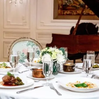 A round table with four plates of gourmet food, glasses of water, and a floral centerpiece, set in an elegant room with a grand piano in the background.