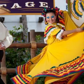 Two dancers perform traditional Mexican folklorico dance on an outdoor stage; the woman wears a bright yellow dress with colorful trim, and the man wears a sombrero and white shirt.