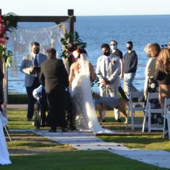 Outdoor wedding ceremony by the water, with guests seated and standing, bride and groom at the altar, and a dog present near the couple.