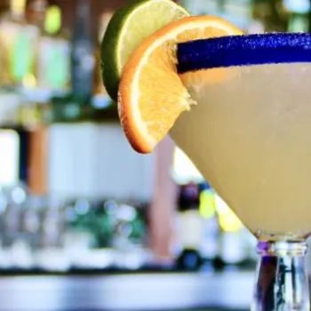 A margarita in a large glass with a blue rim, garnished with orange and lime slices, sits on a bar counter with blurred bottles in the background.
