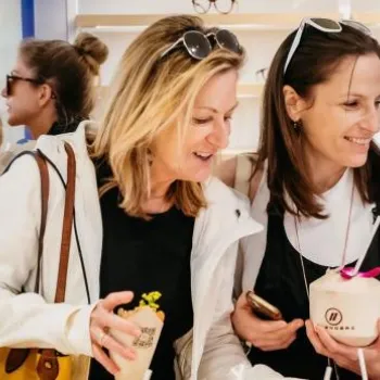 Two women smile and hold drinks at an indoor event, with a "Grand Opening March 11, 2023" sign visible in the background. Several people are gathered around them.