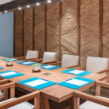 A modern conference room with a wooden table, white chairs, blue folders on each seat, a mounted TV, decorative plants, and a surfboard against a textured wooden wall.