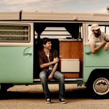Three men with casual attire pose around a vintage mint-green van parked outdoors near trees.