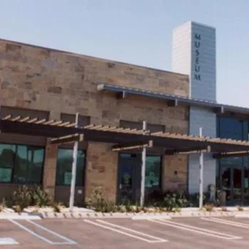 A modern one-story museum building with a stone facade, large windows, an overhang, and an American flag in front, next to an empty parking lot.