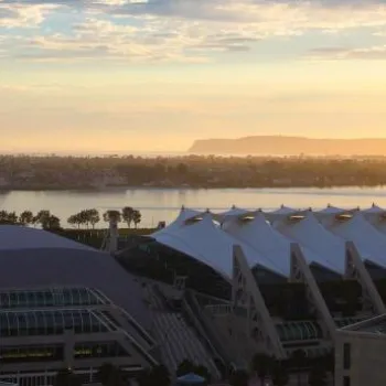 A view of the San Diego Convention Center with its distinctive white sails at sunset, overlooking the bay and distant landforms under a partly cloudy sky.