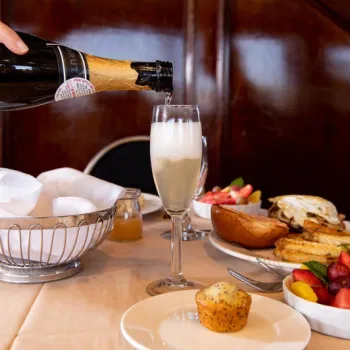A hand pours sparkling wine into a glass on a table set with breakfast foods, including toast, eggs, fruit, and a muffin.