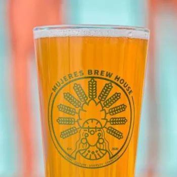A pint glass filled with golden beer, featuring the Mujeres Brew House logo, set against a background of orange and blue stripes.