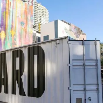 Outdoor urban venue with “QUARTYARD” painted on a white shipping container, colorful murals on nearby buildings, and seating area with string lights.