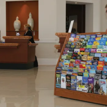 A brochure rack filled with pamphlets stands in the lobby of a building, with three people at reception desks in the background.