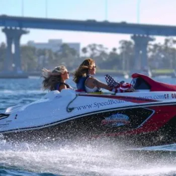 Two people ride a small speed boat on a body of water with a bridge and trees visible in the background.