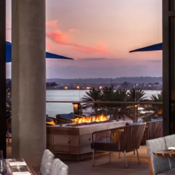 Modern restaurant interior with set tables, large windows, and a view of an outdoor fire pit, patio seating, and water at sunset.