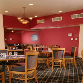 A restaurant dining area with empty wooden tables and chairs, set with glasses and silverware, decorated with framed posters on red and beige walls.