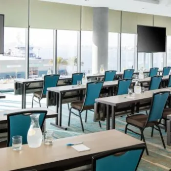 A modern conference room with rows of tables and teal chairs, each set with water pitchers, glasses, and notepads; large windows and two wall-mounted screens are visible.