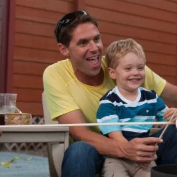 Three people sit outdoors roasting marshmallows over a fire pit, with a child watching and smiling. Houses and trees are visible in the background.