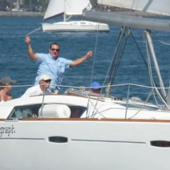 A group of people sits and stands on a white sailboat named "Coconut Telegraph" on calm blue water, some raising their arms in celebration.
