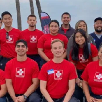 A group of people, some in red shirts with a white cross symbol, pose outdoors in front of banners and palm trees, smiling at the camera.