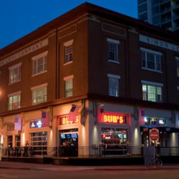 A brick corner building at dusk with neon signs reading "Bub's" and "Simon Levi Company," featuring large windows and a lit outdoor patio.