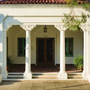 A white, single-story building with a tile roof, columned entrance, green-trimmed windows, and two large potted plants flanking the doorway.