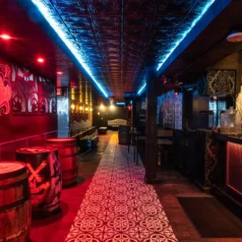 A dimly lit bar with red and blue lighting, wooden barrels as tables, patterned floor tiles, and a mural of people on the wall behind the bar.
