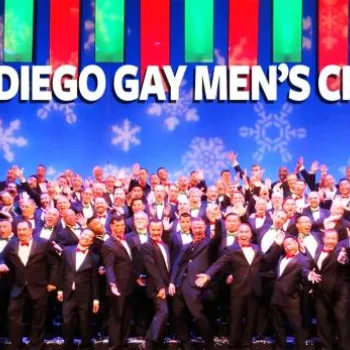 A large group of men in tuxedos pose on stage under “San Diego Gay Men’s Chorus” text, with a colorful, festive background featuring snowflakes and vertical light panels.