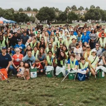 Large group of people posing outdoors on a grassy field, many wearing safety vests and holding cleanup tools and buckets, with tents and trees in the background.