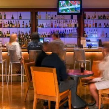 Several people sit at tables and barstools inside a modern bar with a well-lit counter, large liquor display, and television screens above the bar.