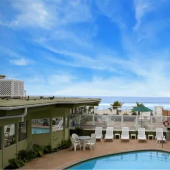 Outdoor swimming pool surrounded by lounge chairs and modern hotel buildings, with a clear blue sky and ocean visible in the background.