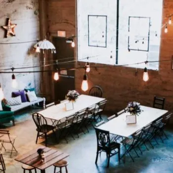 Industrial-style cafe interior with wooden tables, string lights, large windows, and a green couch along a concrete wall.