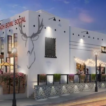 A modern white building labeled "Punky Bowl Social" with outdoor seating, string lights, and palm trees, seen at dusk with people nearby and cars passing by.