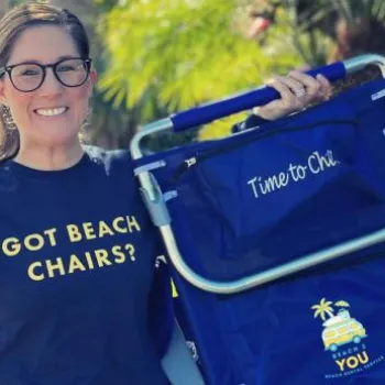 A woman wearing glasses and a "Got Beach Chairs?" sweatshirt holds a blue folding beach chair with "Time to Chill" printed on it. Palm trees are visible in the background.