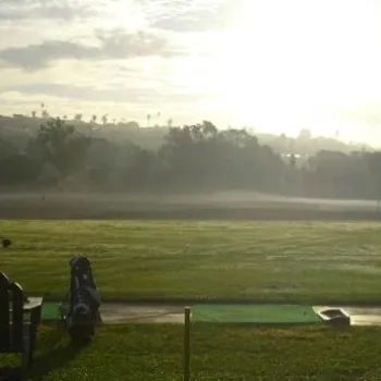 A person practices golf at a driving range at sunrise, with mist over the field and trees in the background.