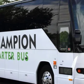 A white charter bus with "Champion Charter Bus" logo is parked outdoors on a paved surface, surrounded by trees.